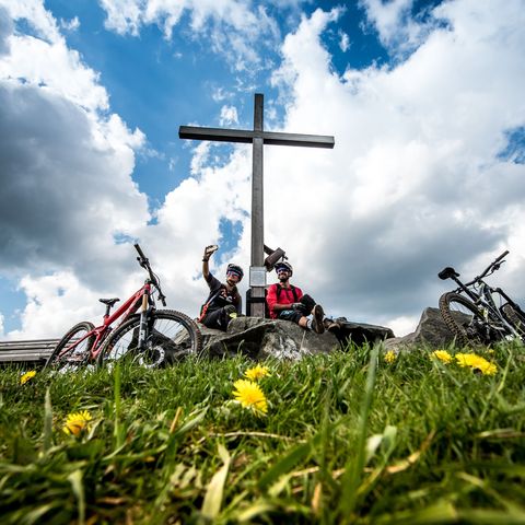 2 Mountainbiker in der Bikewelt Willingen am Gipfelkreuz vor blauem Himmel mit Wolken