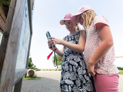 barrierefreien BildbeschreibungstextZwei junge Mädchen mit rosa Kappen stehen nebeneinander im Profil vor einer großen hölzernen Orientierungstafel im Freien. Das eine Mädchen hält ein Smartphone hoch, um einen Inhalt der Infotafel zu scannen, während das andere aufmerksam zusieht. Im Hintergrund ist ein leicht bewölkter Sommerhimmel über dem Freizeitgelände zu erkennen. Two young girls wearing pink caps stand side by side in profile in front of a large wooden information board outdoors, one scanning with a smartphone.