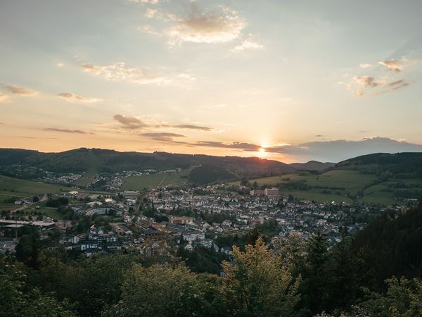 Sonnenuntergang über Willingen im Upland barrierefreien BildbeschreibungstextPanoramaaufnahme von Willingen im Upland bei Sonnenuntergang. Die Sonne geht hinter sanften, bewaldeten Hügeln unter und hüllt den Ort im Tal sowie die umliegenden grünen Wiesen und Wälder in warmes Abendlicht. Im Vordergrund stehen begrünte Baumkronen.barrierefreien BildbeschreibungstextPanoramic view of Willingen nestled in a valley surrounded by wooded hills at sunset. The low sun casts a golden glow across the landscape, fields, and houses, with leafy treetops in the foreground.