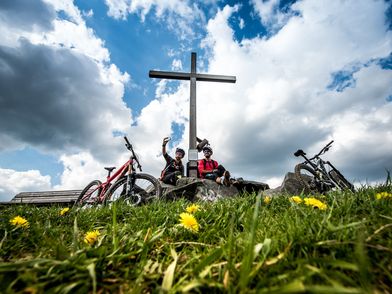 2 Mountainbiker am Gipfelkreuz des Clemensbergs vor beeindruckendem Wolkenhimmel, im Vordergrund Löwenzahn